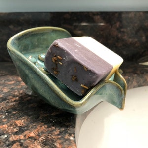 May include: A teal ceramic soap dish with a wavy edge holds a bar of lavender soap with dried herbs. The soap dish is sitting on a granite countertop.