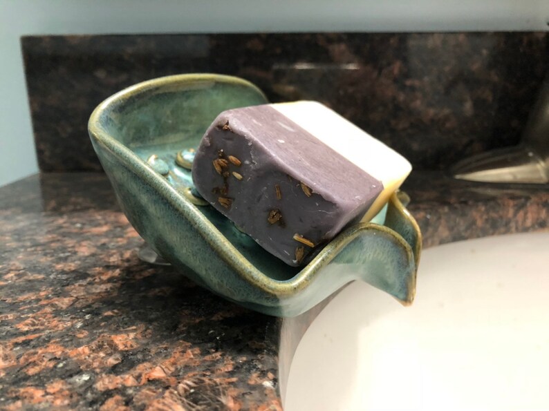 May include: A teal ceramic soap dish with a wavy edge holds a bar of lavender soap with dried herbs. The soap dish is sitting on a granite countertop.