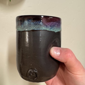 May include: A handmade ceramic mug with a dark brown body and a rim glazed in shades of blue and purple. The mug has a smooth, cylindrical shape and a small, embossed design near the base. The mug is held by a hand.