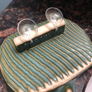May include: A green and white ceramic soap dish with two suction cups on the back. The dish has a striped pattern.