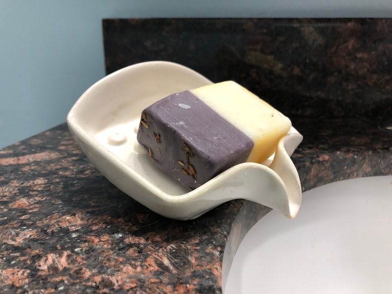 May include: A white ceramic soap dish with a curved design holds a bar of soap. The soap is half purple and half yellow.