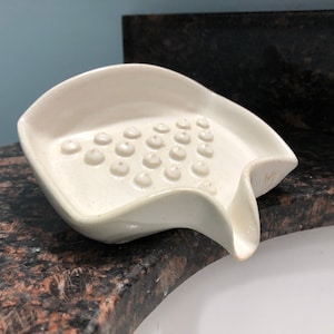 May include: White ceramic soap dish with a textured surface and a drain spout. The dish has a unique shape and is designed to hold soap bars.