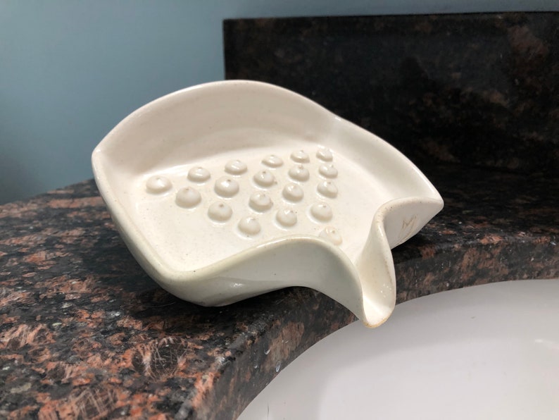 May include: White ceramic soap dish with a textured surface and a drain spout. The dish has a unique shape and is designed to hold soap bars.
