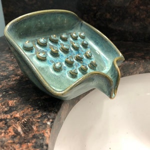May include: A teal blue ceramic soap dish with a textured surface and a drain spout. The dish has a series of small, round indentations on the bottom.