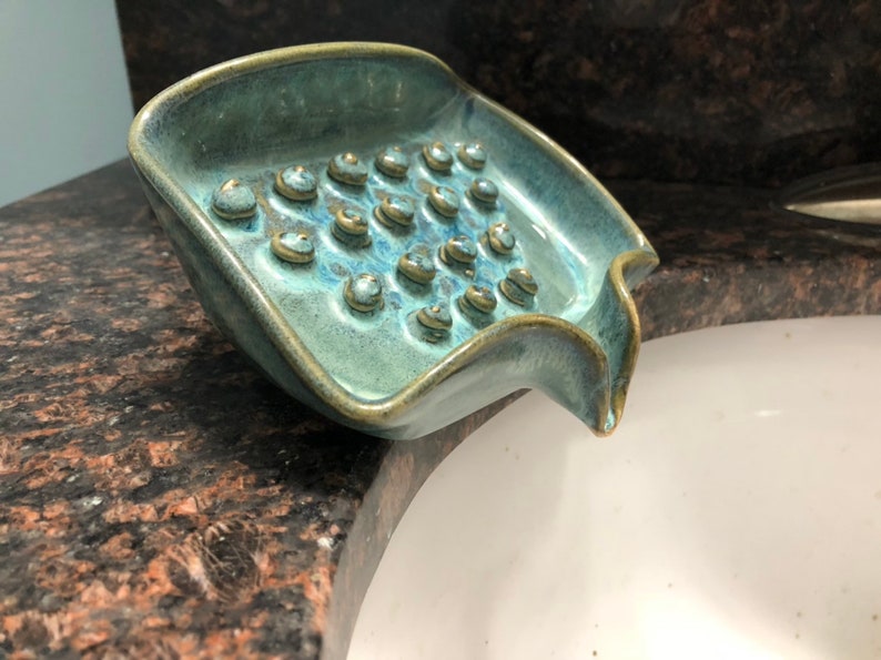 May include: A teal blue ceramic soap dish with a textured surface and a drain spout. The dish has a series of small, round indentations on the bottom.