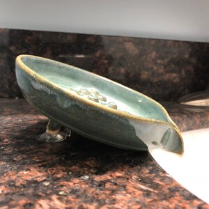 May include: A blue ceramic soap dish with a spout and suction cups on the bottom. The dish is sitting on a countertop and is tilted, with soap suds spilling out of the spout.