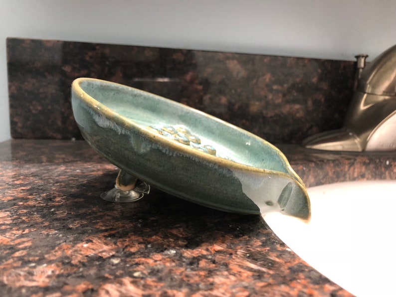 May include: A blue ceramic soap dish with a spout and suction cups on the bottom. The dish is sitting on a countertop and is tilted, with soap suds spilling out of the spout.