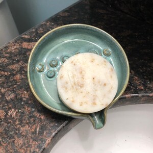 May include: A round, white bar of soap with flecks of brown sits in a teal ceramic soap dish with a spout. The dish has a textured surface with small, raised circles.