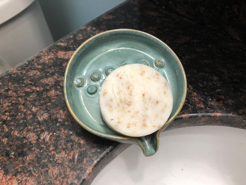May include: A round, white bar of soap with flecks of brown sits in a teal ceramic soap dish with a spout. The dish has a textured surface with small, raised circles.