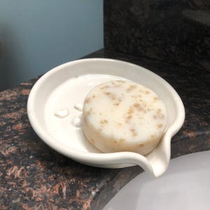 May include: A white ceramic soap dish with a rounded shape and a spout. A white bar of soap with brown flecks is resting in the dish.