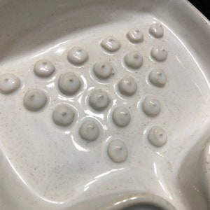 May include: A white ceramic dish with a textured surface and multiple small, round indentations. The dish is designed for holding and drying soap.