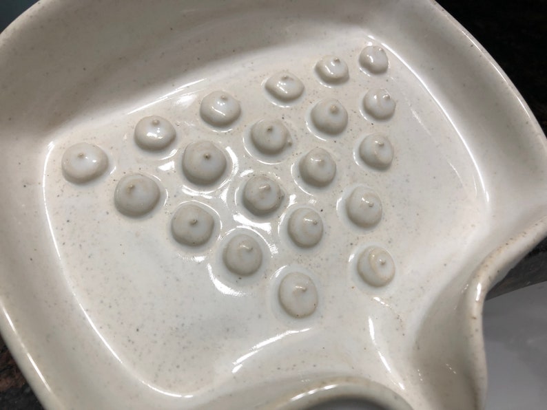 May include: A white ceramic dish with a textured surface and multiple small, round indentations. The dish is designed for holding and drying soap.