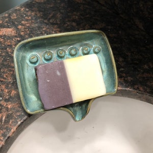 May include: A teal ceramic soap dish with a decorative design and a bar of soap with purple and white stripes.
