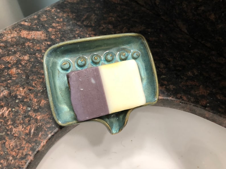May include: A teal ceramic soap dish with a decorative design and a bar of soap with purple and white stripes.