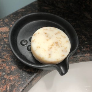 May include: A white bar of soap with flecks of gold in a black soap dish with a spout.
