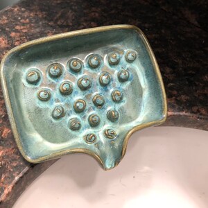 May include: A teal green ceramic soap dish with a textured surface and gold accents. The dish has a unique shape with a curved spout and a series of small, raised circles.