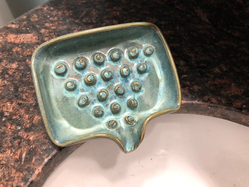 May include: A teal green ceramic soap dish with a textured surface and gold accents. The dish has a unique shape with a curved spout and a series of small, raised circles.