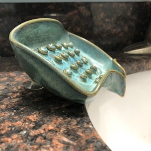 May include: A teal blue ceramic soap dish with a textured surface and a built-in drainage spout. The dish has a suction cup on the bottom to keep it in place.