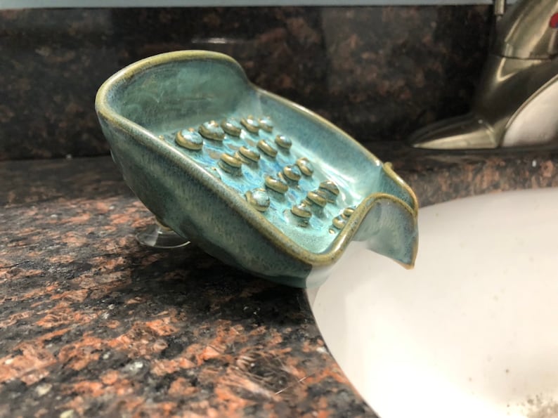 May include: A teal blue ceramic soap dish with a textured surface and a built-in drainage spout. The dish has a suction cup on the bottom to keep it in place.