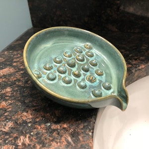 May include: A teal green ceramic soap dish with a textured surface and a drain spout. The dish has a pattern of small, raised circles.