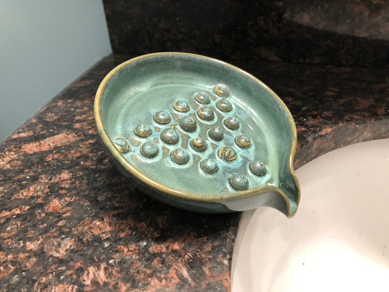 May include: A teal green ceramic soap dish with a textured surface and a drain spout. The dish has a pattern of small, raised circles.
