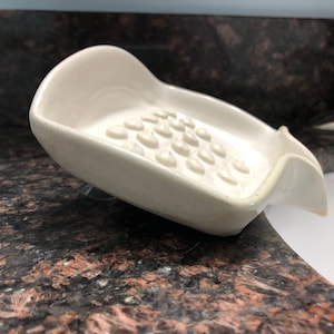 May include: White ceramic soap dish with a textured surface and a curved design. The dish is designed to hold a bar of soap and drain excess water.