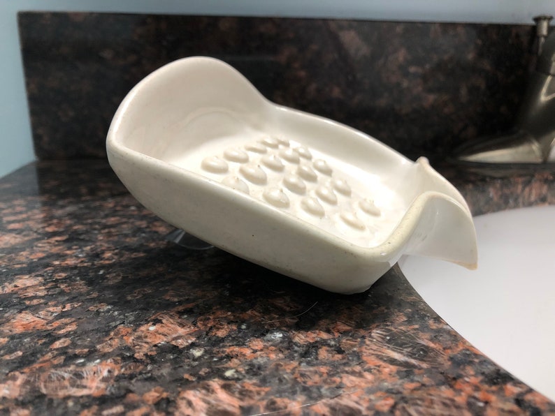 May include: White ceramic soap dish with a textured surface and a curved design. The dish is designed to hold a bar of soap and drain excess water.