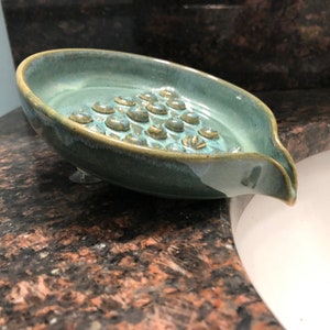 May include: A teal blue ceramic soap dish with a textured bottom and a spout for draining water. The dish has suction cups on the bottom to keep it in place.