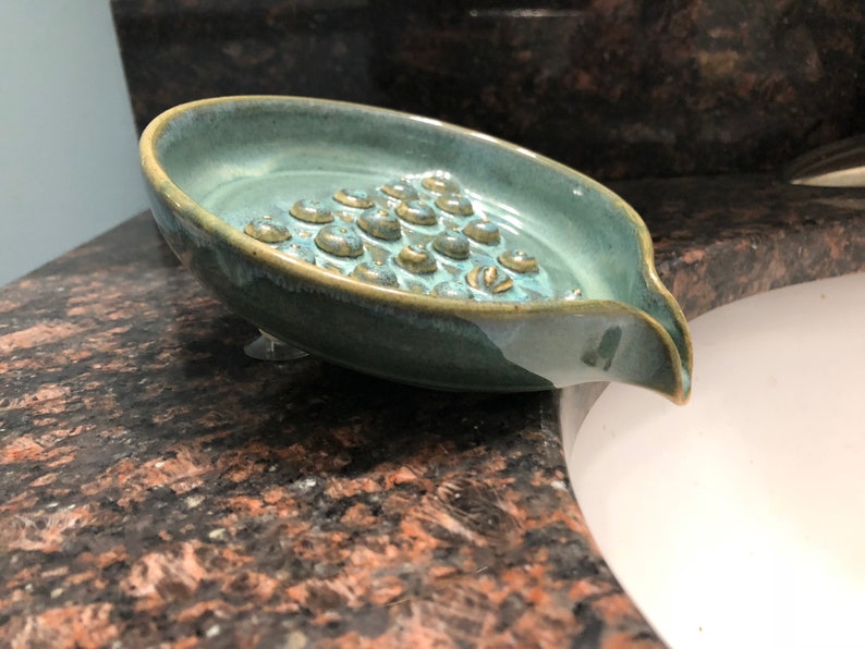 May include: A teal blue ceramic soap dish with a textured bottom and a spout for draining water. The dish has suction cups on the bottom to keep it in place.