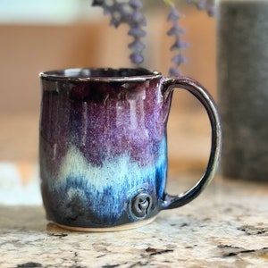 Może przedstawiać: Ręcznie robiony kubek ceramiczny z fioletową, niebieską i białą glazurą. Kubek ma zaokrąglone dno i uchwyt. Kubek ma małe, okrągłe logo z boku.