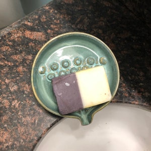 May include: A teal ceramic soap dish with a raised design of circles. A bar of soap with a purple and white design is resting in the dish.