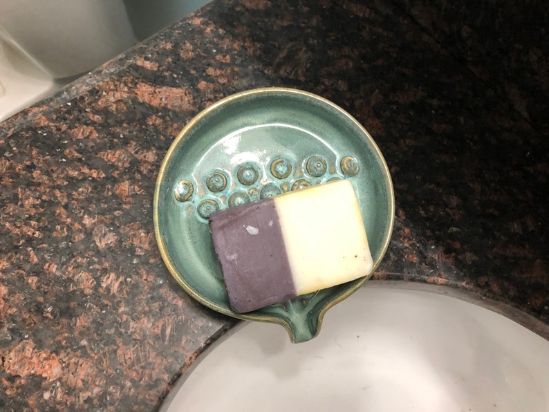 May include: A teal ceramic soap dish with a raised design of circles. A bar of soap with a purple and white design is resting in the dish.