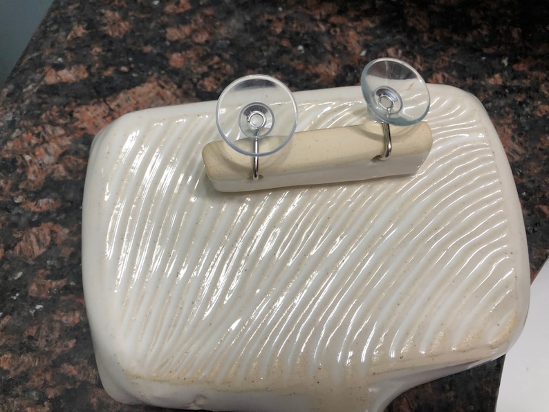 May include: White ceramic dish with a textured surface. Two suction cups with hooks are attached to a wooden bar, which is placed on top of the dish.