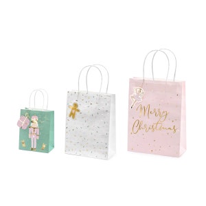 May include: Three festive gift bags with white handles. One is mint green with a nutcracker and gift graphic. The second is white with gold confetti and a gingerbread man. The third is pink with "Merry Christmas" text and a ballerina.