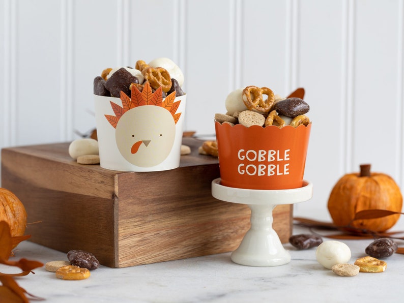 K&ouml;nnte beinhalten: Zwei Papierbecher gef&uuml;llt mit einer Mischung aus Brezeln, Schokoladenbonbons und wei&szlig;en Schokoladenst&uuml;ckchen. Ein Becher ist mit einem Truthahn-Design und dem Text "Gobble Gobble" verziert. Der andere Becher ist mit einem Truthahn-Design und dem Text "Gobble Gobble" verziert.