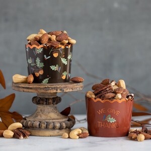 K&ouml;nnte beinhalten: Zwei kleine Becher gef&uuml;llt mit gemischten N&uuml;ssen, einer mit einem schwarzen und braunen Herbstdesign und der andere mit dem Text "We Give Thanks". Die Becher stehen auf einem Holzsockel vor einem grauen Hintergrund.