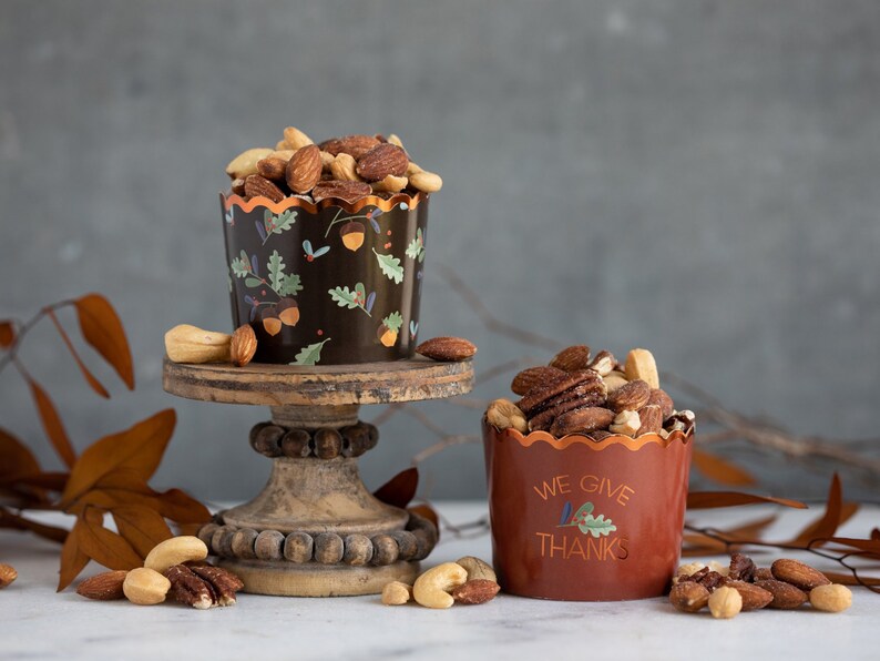 K&ouml;nnte beinhalten: Zwei kleine Becher gef&uuml;llt mit gemischten N&uuml;ssen, einer mit einem schwarzen und braunen Herbstdesign und der andere mit dem Text "We Give Thanks". Die Becher stehen auf einem Holzsockel vor einem grauen Hintergrund.