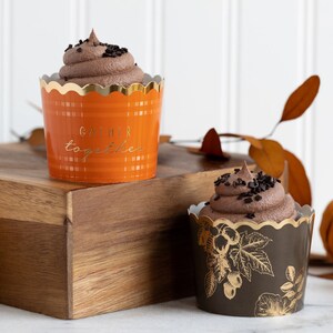 K&ouml;nnte beinhalten: Zwei Schokoladen-Cupcakes mit Schokoladenstreuseln. Der Cupcake links ist in einer orangefarbenen und goldenen Cupcake-Form mit dem Text "Gather together". Der Cupcake rechts ist in einer schwarzen und goldenen Cupcake-Form mit einem Blumenmuster.