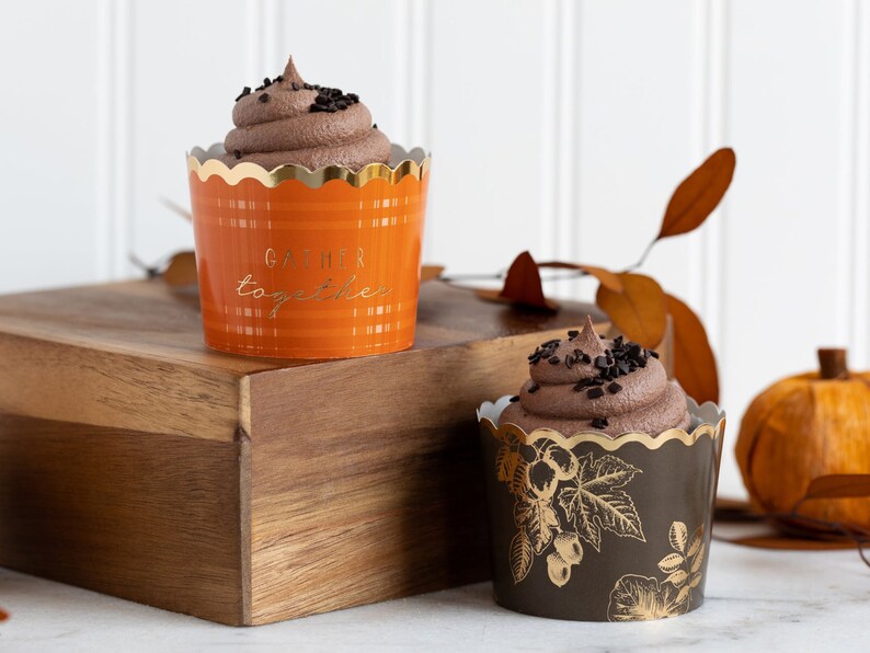 K&ouml;nnte beinhalten: Zwei Schokoladen-Cupcakes mit Schokoladenstreuseln. Der Cupcake links ist in einer orangefarbenen und goldenen Cupcake-Form mit dem Text "Gather together". Der Cupcake rechts ist in einer schwarzen und goldenen Cupcake-Form mit einem Blumenmuster.