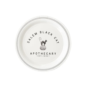 May include: White paper plate with a black rim and the words "Salem Black Cat Apothecary Est. 1693" printed in black. A black cat is perched on a pumpkin in the center of the plate.