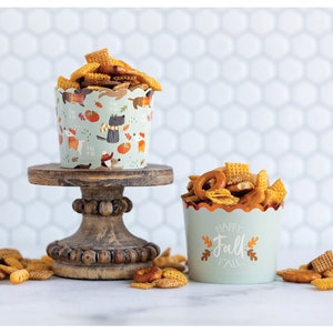 K&ouml;nnte beinhalten: Zwei Papierbecher gef&uuml;llt mit einem Snack-Mix aus Brezeln, M&uuml;sliquadraten und anderen knusprigen Snacks. Der Becher links hat ein Herbst-Design mit einer schwarzen Katze, K&uuml;rbissen und Bl&auml;ttern. Der Becher rechts hat einen gr&uuml;nen Hintergrund mit dem Text "Happy Fall" in Wei&szlig; und Orange.