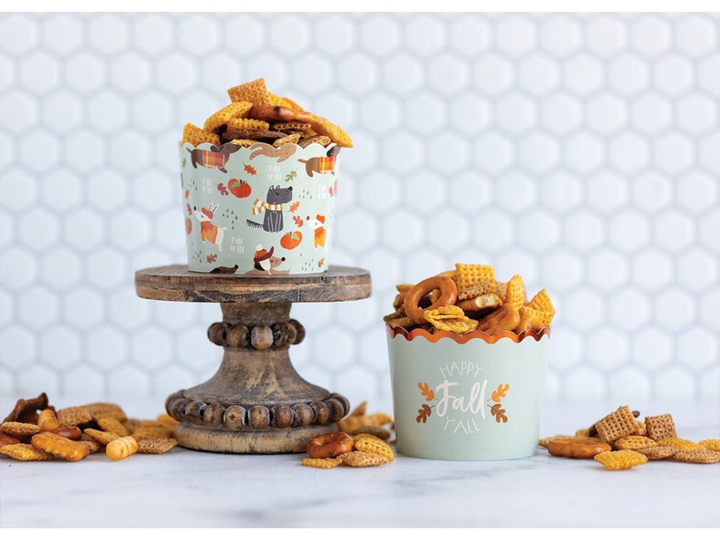 K&ouml;nnte beinhalten: Zwei Papierbecher gef&uuml;llt mit einem Snack-Mix aus Brezeln, M&uuml;sliquadraten und anderen knusprigen Snacks. Der Becher links hat ein Herbst-Design mit einer schwarzen Katze, K&uuml;rbissen und Bl&auml;ttern. Der Becher rechts hat einen gr&uuml;nen Hintergrund mit dem Text "Happy Fall" in Wei&szlig; und Orange.