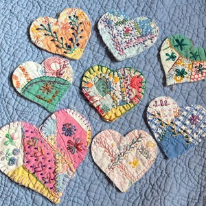 May include: A collection of nine handmade heart-shaped fabric patches. Each heart features a unique patchwork design with embroidered details in various colors, including pink, yellow, blue, and green. The hearts are arranged on a light blue quilted background.