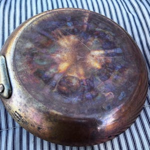 May include: A well-used copper-bottomed frying pan with a long handle. The pan's underside displays a spectrum of colors, including bronze, purple, and gold, indicating heat exposure. The handle is riveted to the pan's body.