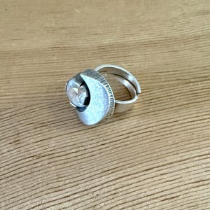 May include: A silver-toned ring featuring a crescent-shaped metal component with a textured, lined edge and a smooth, matte finish. A single, clear, faceted round stone is set within the curve of the crescent, attached to a simple, open-ended band.