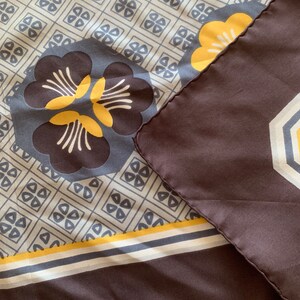 May include: A brown silk scarf with a geometric pattern in shades of yellow, white, and gray. The scarf features a large floral design in the center.
