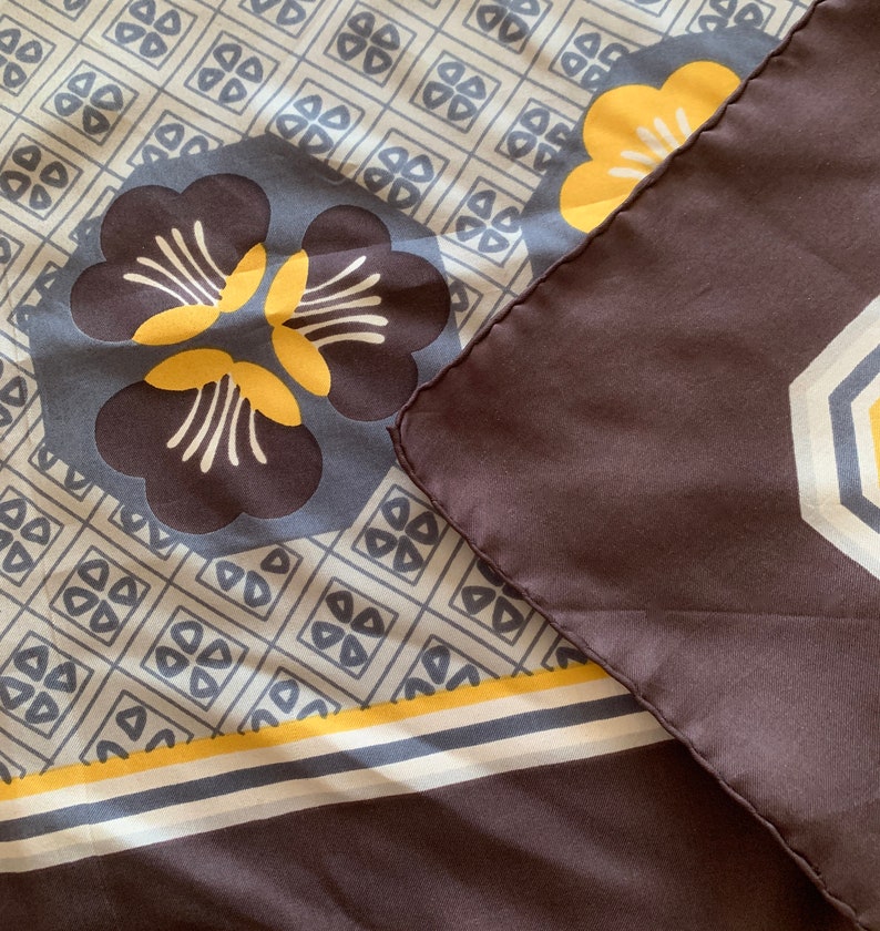 May include: A brown silk scarf with a geometric pattern in shades of yellow, white, and gray. The scarf features a large floral design in the center.
