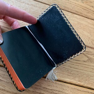 May include: Open black notebook with gold leaf trim and orange-edged pages. A white ribbon bookmark with a tassel is visible. The notebook is held open, revealing the blank pages.