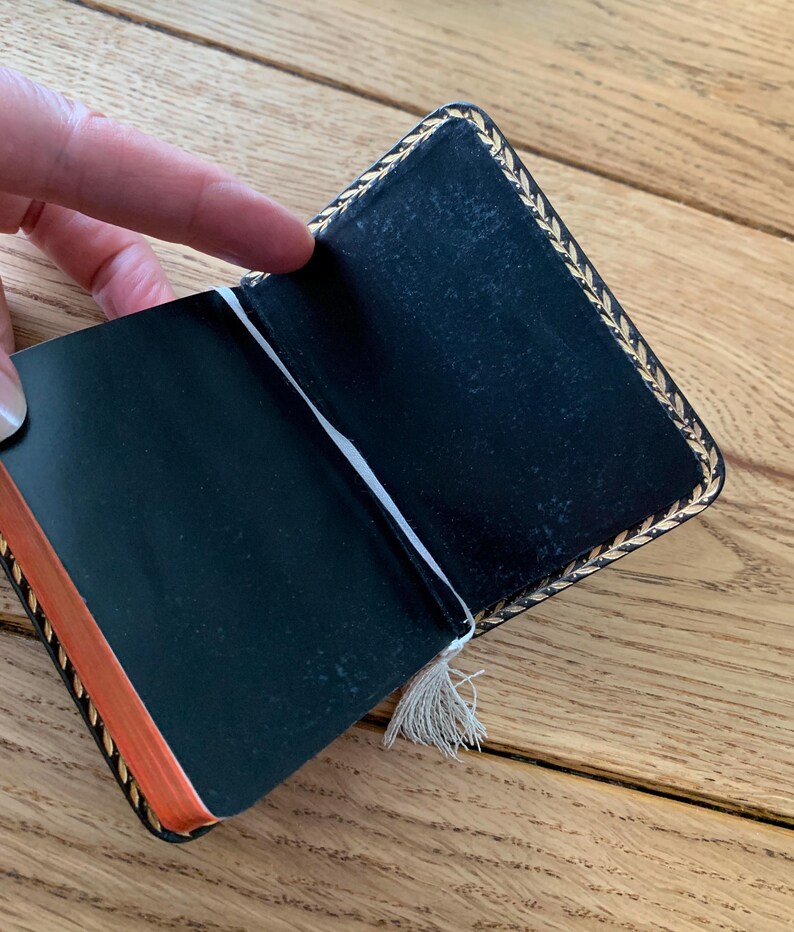 May include: Open black notebook with gold leaf trim and orange-edged pages. A white ribbon bookmark with a tassel is visible. The notebook is held open, revealing the blank pages.