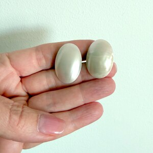 May include: A pair of oval-shaped earrings with a smooth, iridescent white finish. The earrings feature a polished, reflective surface that catches the light, showcasing a subtle pearlescent sheen across the curved, rounded edges of each piece.